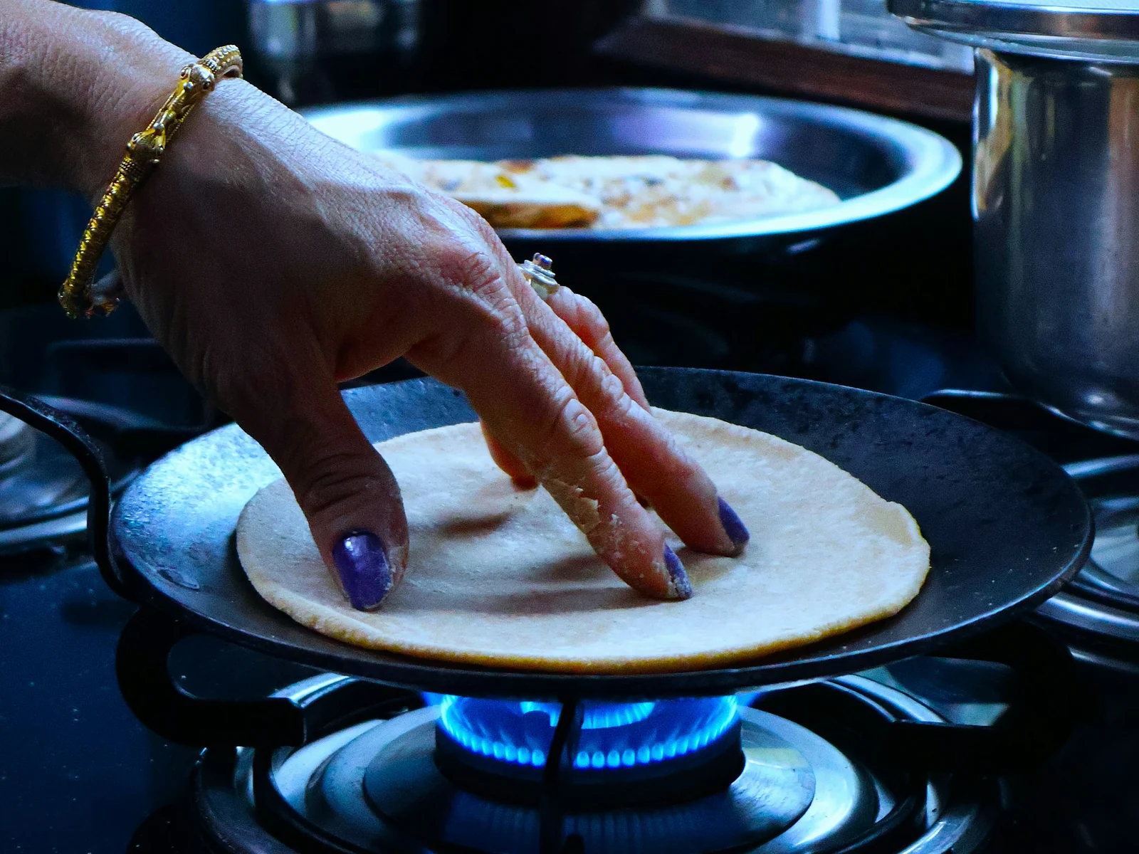 A hand pressing a roti on a hot tawa over a gas flame in a home kitchen